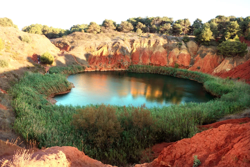 Ancienne carrière de bauxite d'Otrante avec parois rouges vives, lac émeraude au fond du cratère et couronne de pins parasols, Salento, Pouilles, Italie